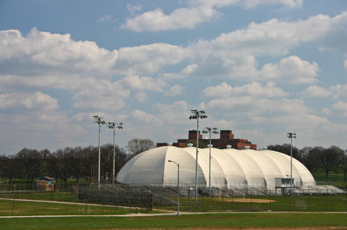 Large white dome sports facility beside a fenced field under a cloudy blue sky.