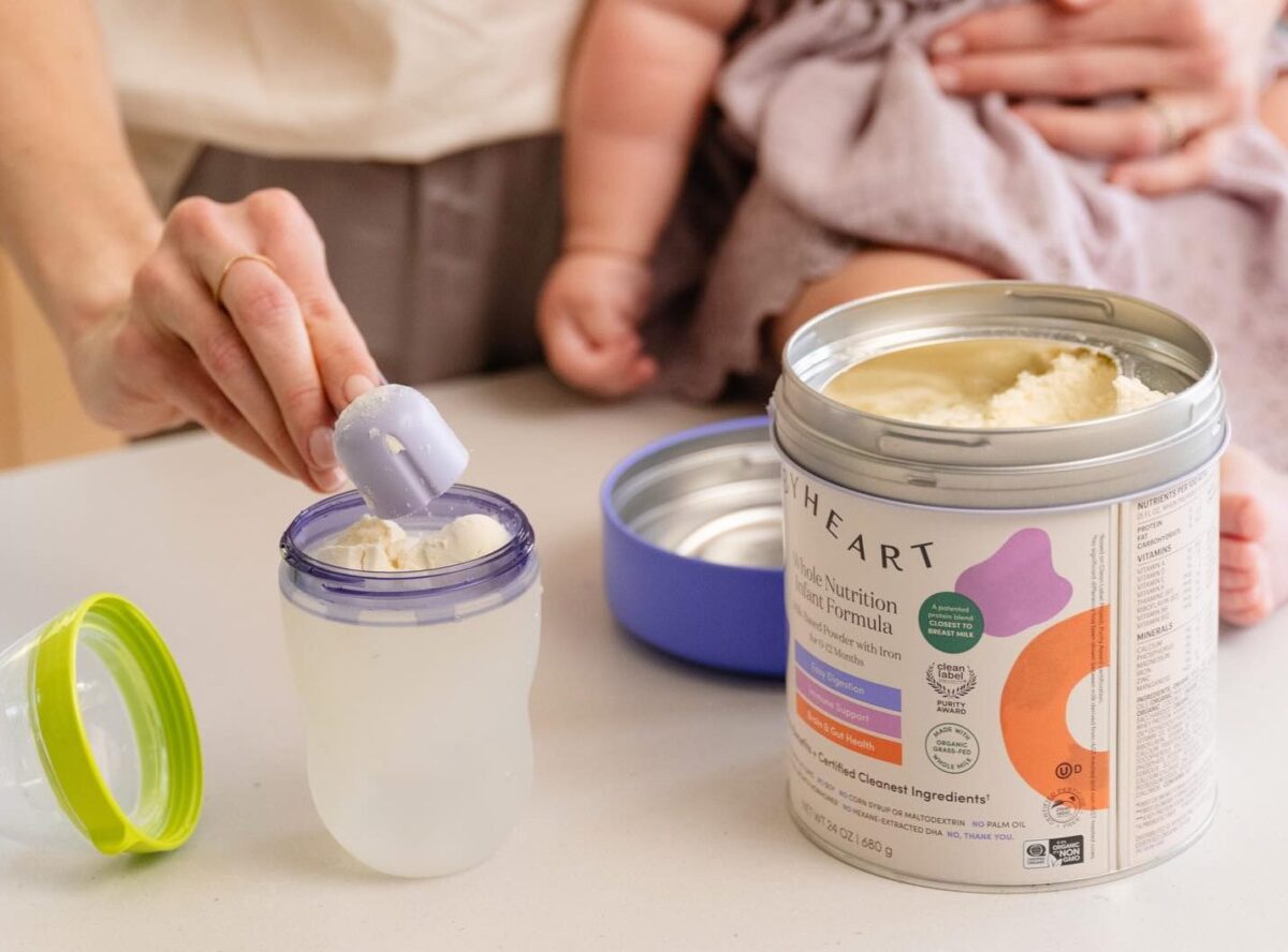 Hand scooping baby formula into a bottle beside an open can with a baby in the background.