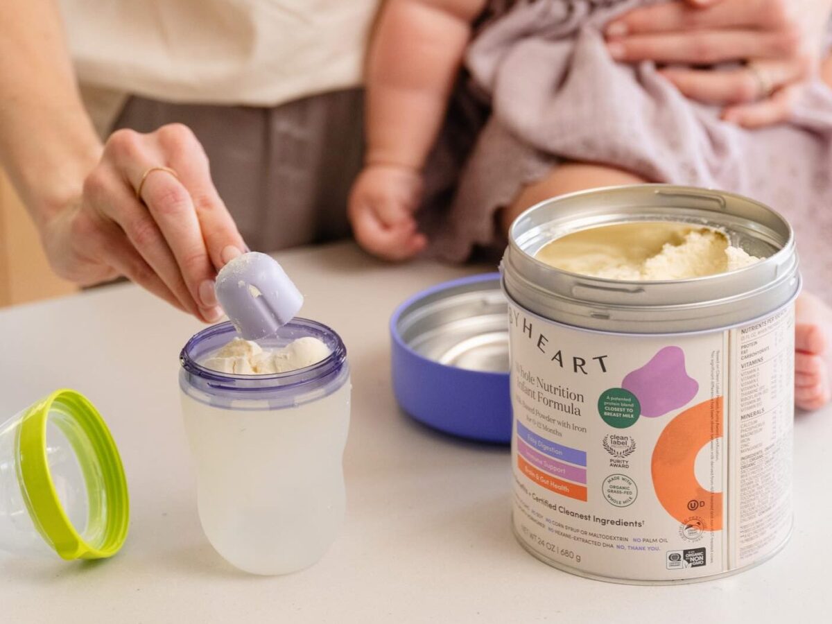 Hand scooping baby formula into a bottle beside an open can with a baby in the background.