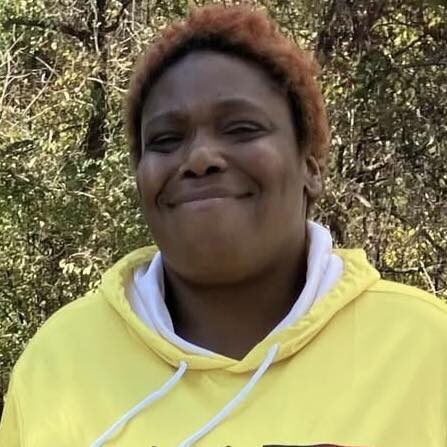 Smiling person in a yellow hoodie standing outdoors with trees behind them.