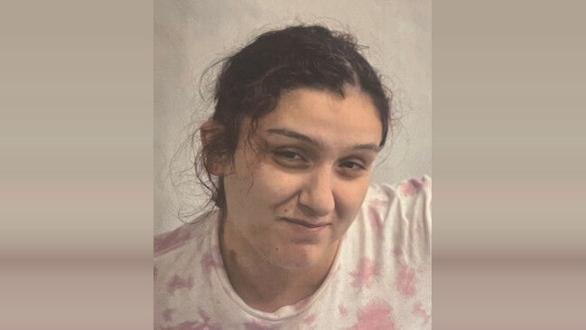 Smiling woman with dark hair in a white and pink tie-dye shirt against a beige wall.