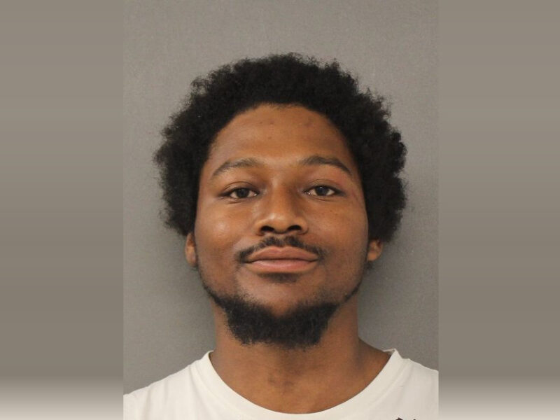 Mugshot-style portrait of a man against a plain gray background wearing a white shirt.