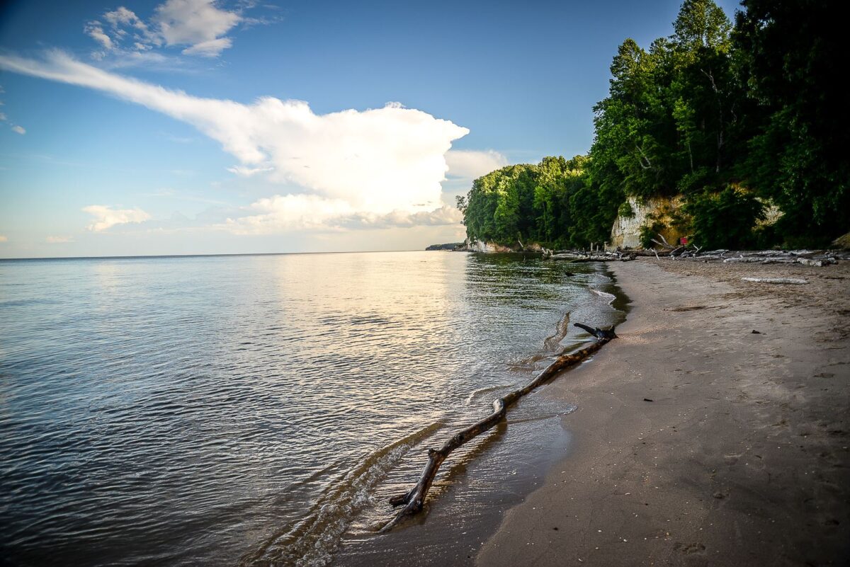 Sandy tropical beach beside calm water and a forested cliff under a blue sky with clouds.