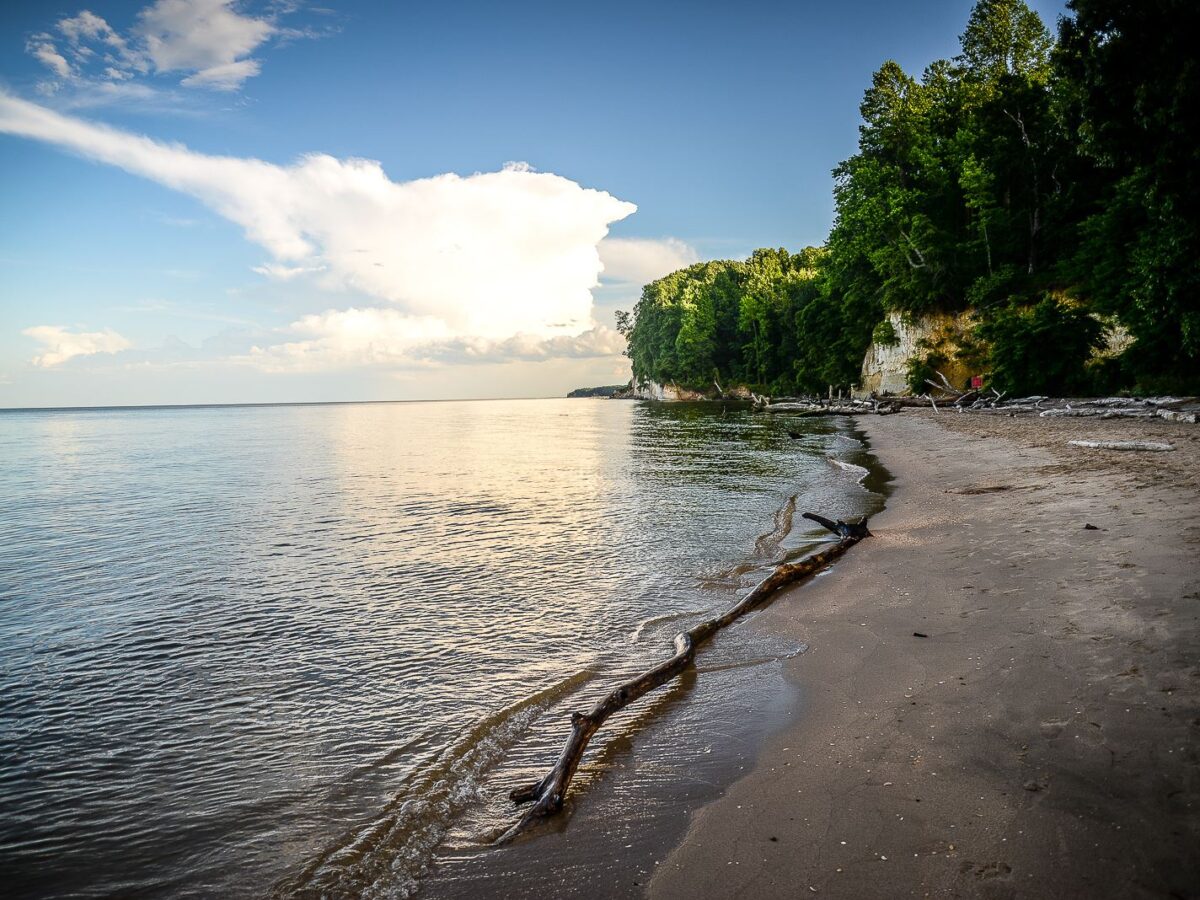 Sandy tropical beach beside calm water and a forested cliff under a blue sky with clouds.