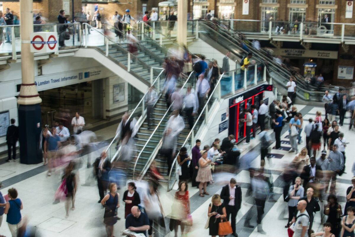 Crowded indoor train station concourse with blurred commuters on escalators and stairs.