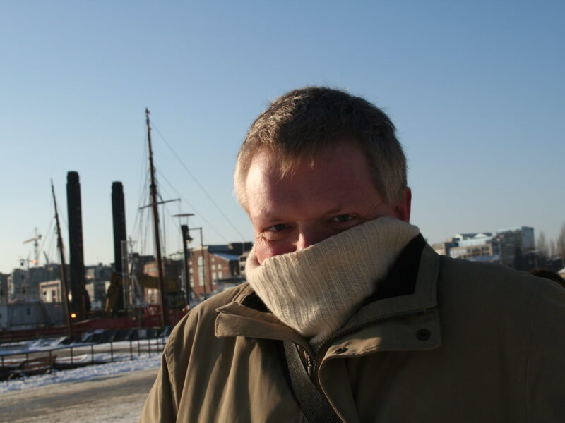 Man in a winter coat and scarf standing outdoors by a harbor on a cold day