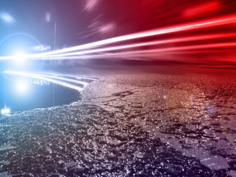 Blue and red light streaks race over a wet road at night with a bright glare on the left.