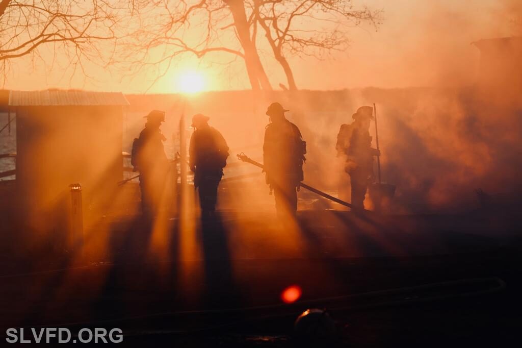 Firefighters silhouetted in smoky orange light at sunset near a burning structure.