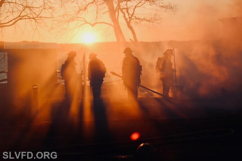 Firefighters silhouetted in smoky orange light at sunset near a burning structure.