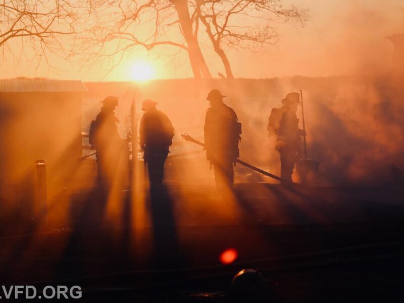 Firefighters silhouetted in smoky orange light at sunset near a burning structure.