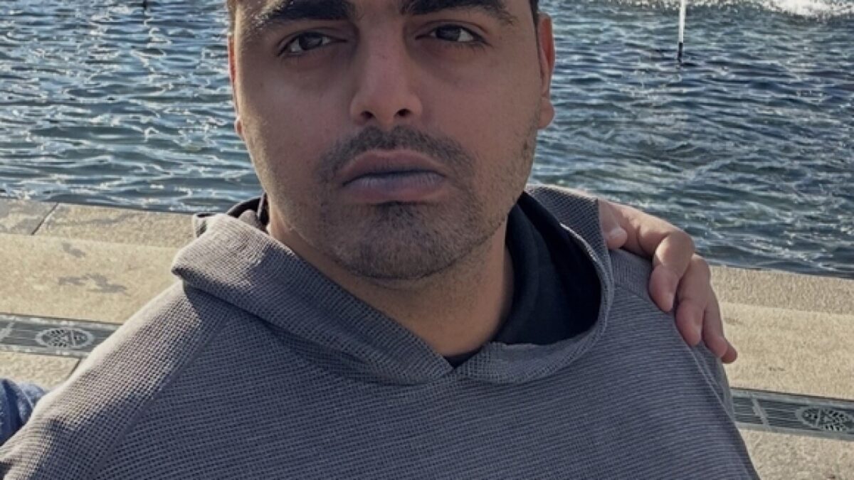 Close-up selfie of a man in a gray hoodie by the water with an arm around his shoulder.