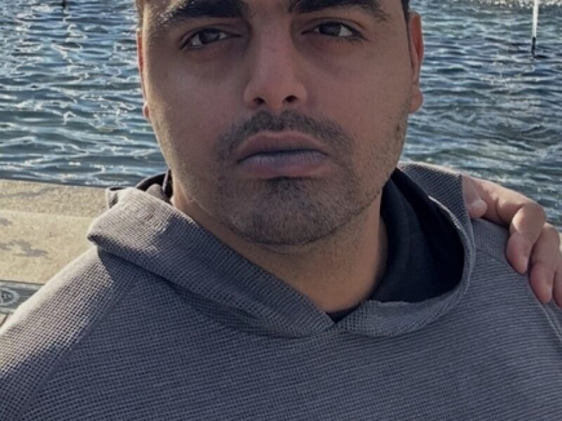 Close-up selfie of a man in a gray hoodie by the water with an arm around his shoulder.