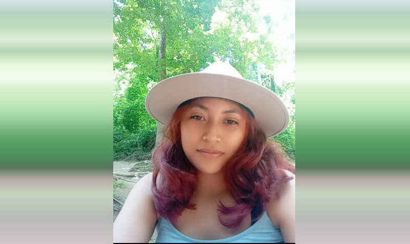 Woman in a wide-brim hat taking a selfie outdoors in a lush green setting.