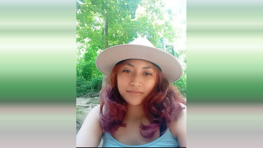 Woman in a wide-brim hat taking a selfie outdoors in a lush green setting.