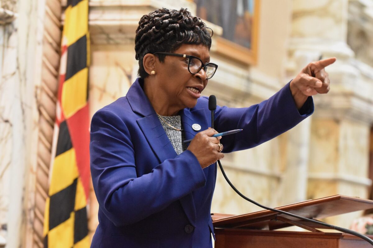 Woman in a purple blazer speaks at a podium and points while holding a microphone.