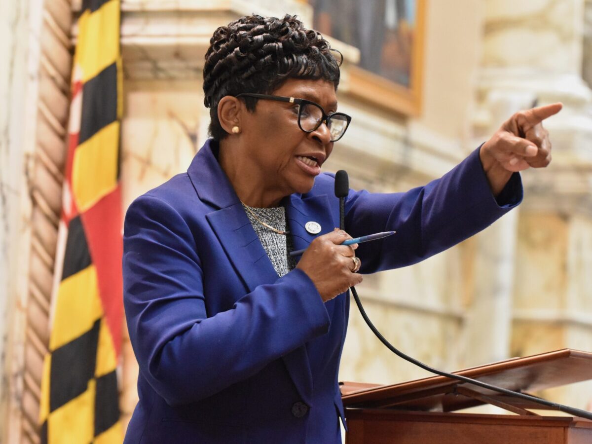 Woman in a purple blazer speaks at a podium and points while holding a microphone.