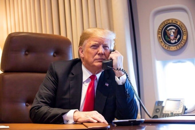 Donald Trump sits at a desk on a phone in a wood-paneled aircraft cabin.