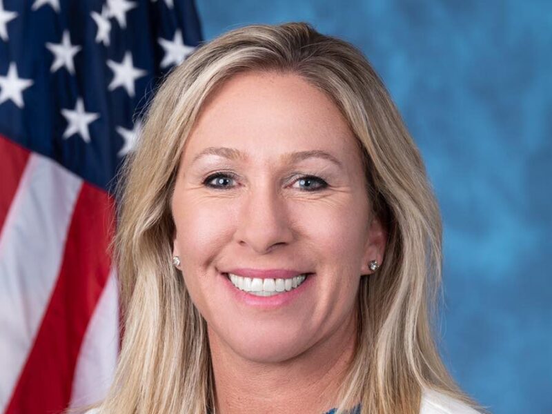 Smiling blonde woman in front of an American flag and blue backdrop.