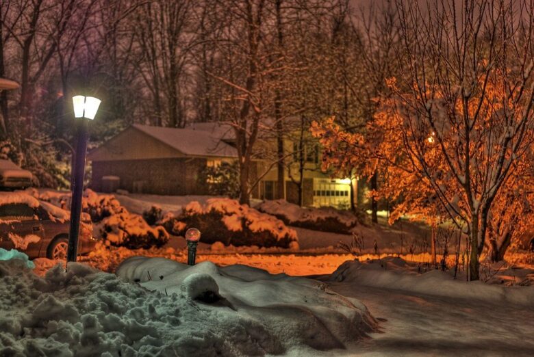 Snowy suburban street at night with glowing lamp posts, lit houses, and orange winter trees.