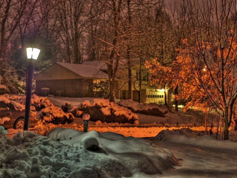 Snowy suburban street at night with glowing lamp posts, lit houses, and orange winter trees.