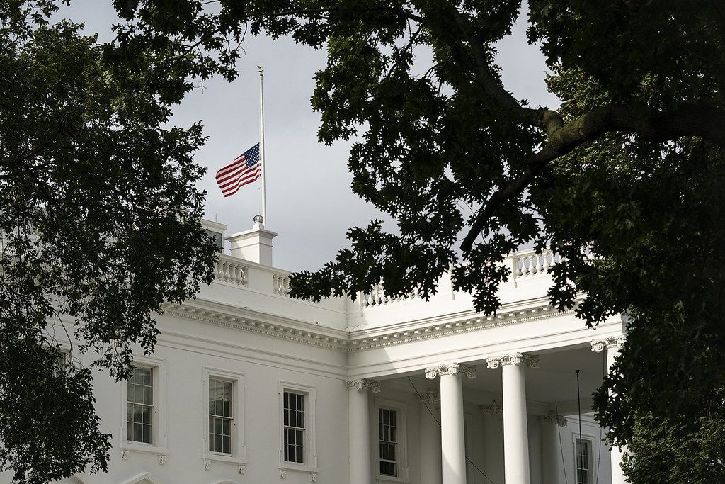 White House exterior framed by tree branches with an American flag flying above the roof.