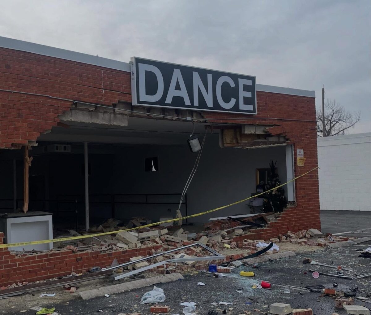 Damaged brick storefront with a large hole beneath a DANCE sign and debris scattered outside.