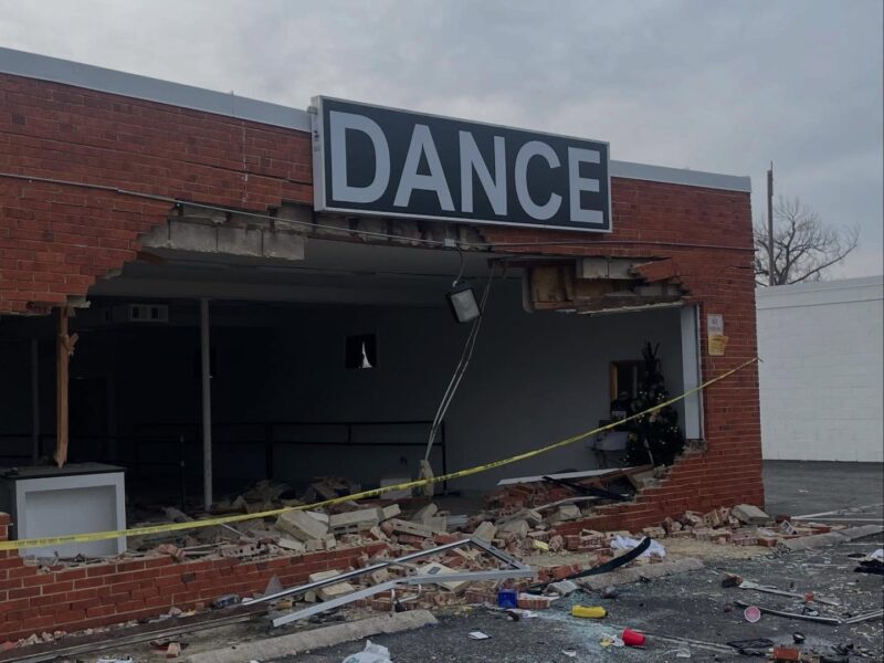 Damaged brick storefront with a large hole beneath a DANCE sign and debris scattered outside.