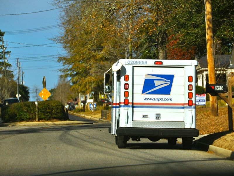 USPS mail truck driving down a quiet suburban street in autumn.