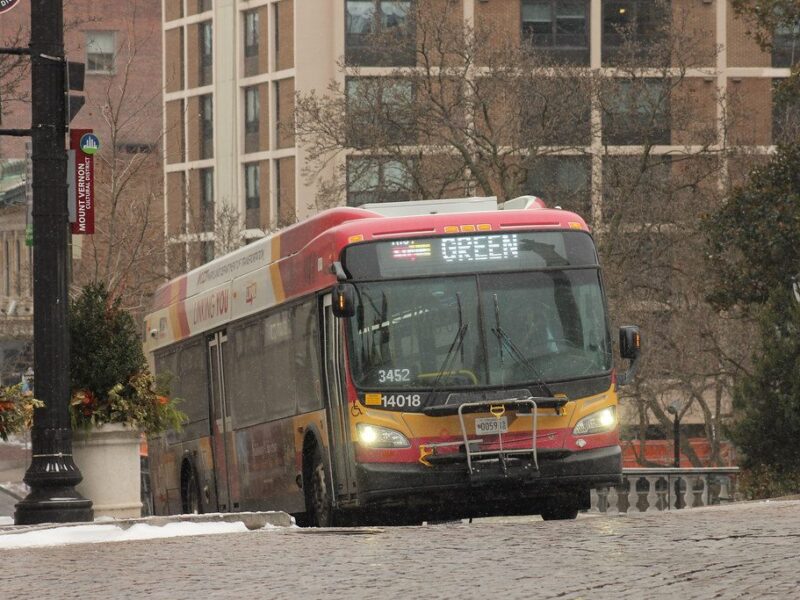 City bus driving up a snowy cobblestone street in an urban neighborhood.