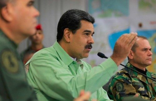 Man in a green shirt speaks into a microphone with a raised hand beside military officers.