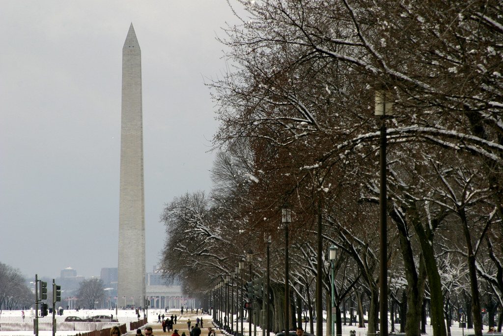 Washington Monument rises beyond a snowy, tree-lined park path under a gray winter sky.