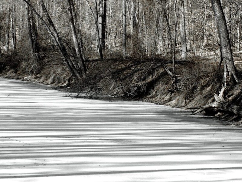 Frozen river bordered by leafless trees with striped shadows across the ice.