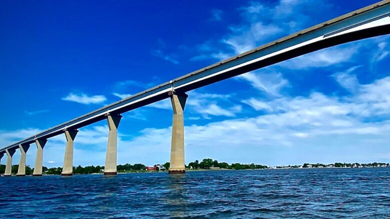 Long concrete bridge stretches across blue water under a bright sky with scattered clouds.