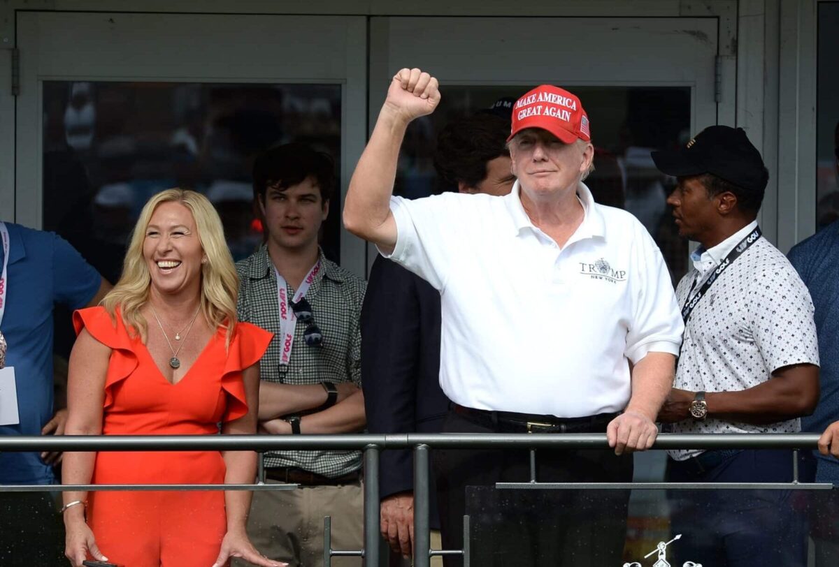 Man in a red cap raises his fist beside a smiling woman in an orange dress on a balcony.