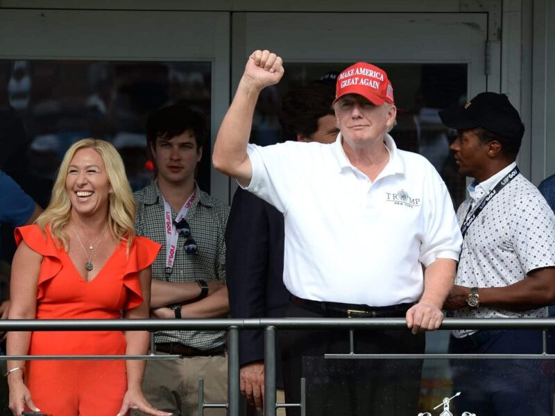 Man in a red cap raises his fist beside a smiling woman in an orange dress on a balcony.