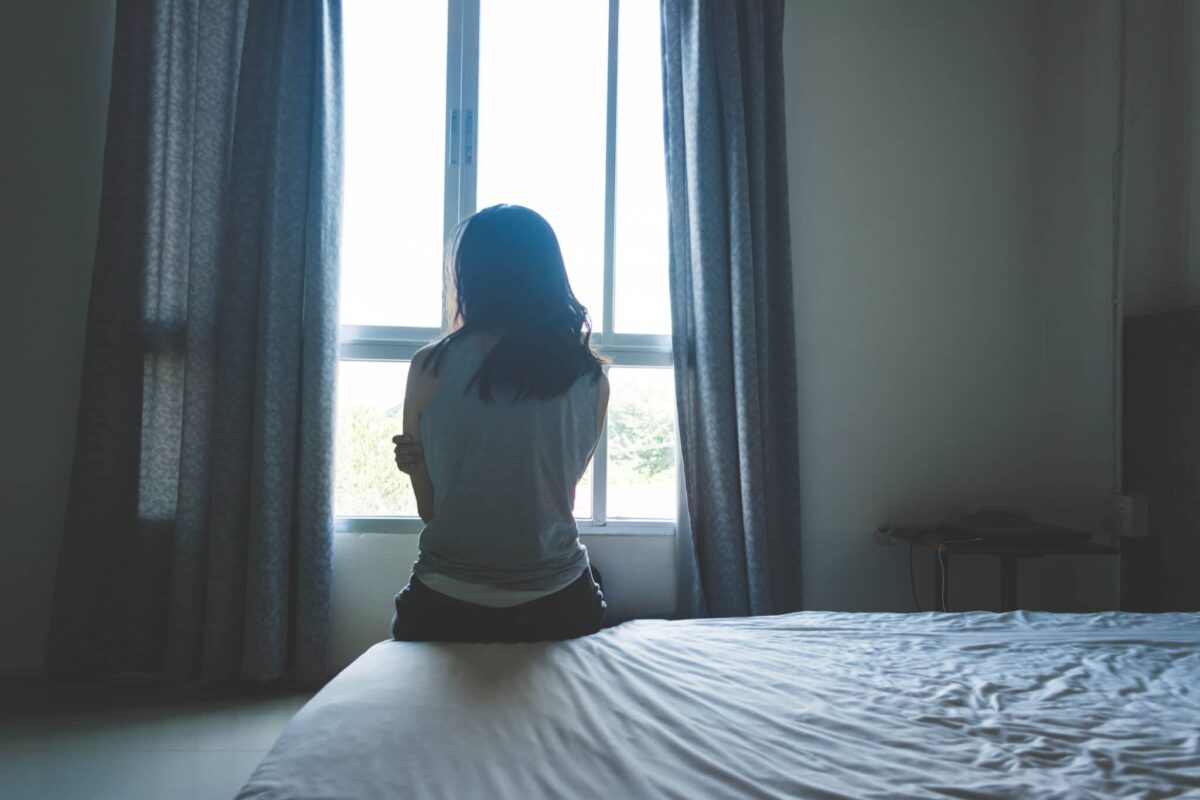 Silhouetted person sitting on a bed facing a bright window with curtains.