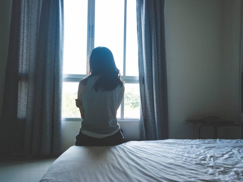 Silhouetted person sitting on a bed facing a bright window with curtains.
