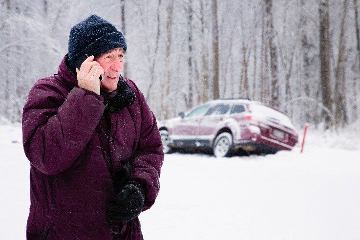 Man in winter coat talking on a phone beside a snow-covered car in a snowy forest.