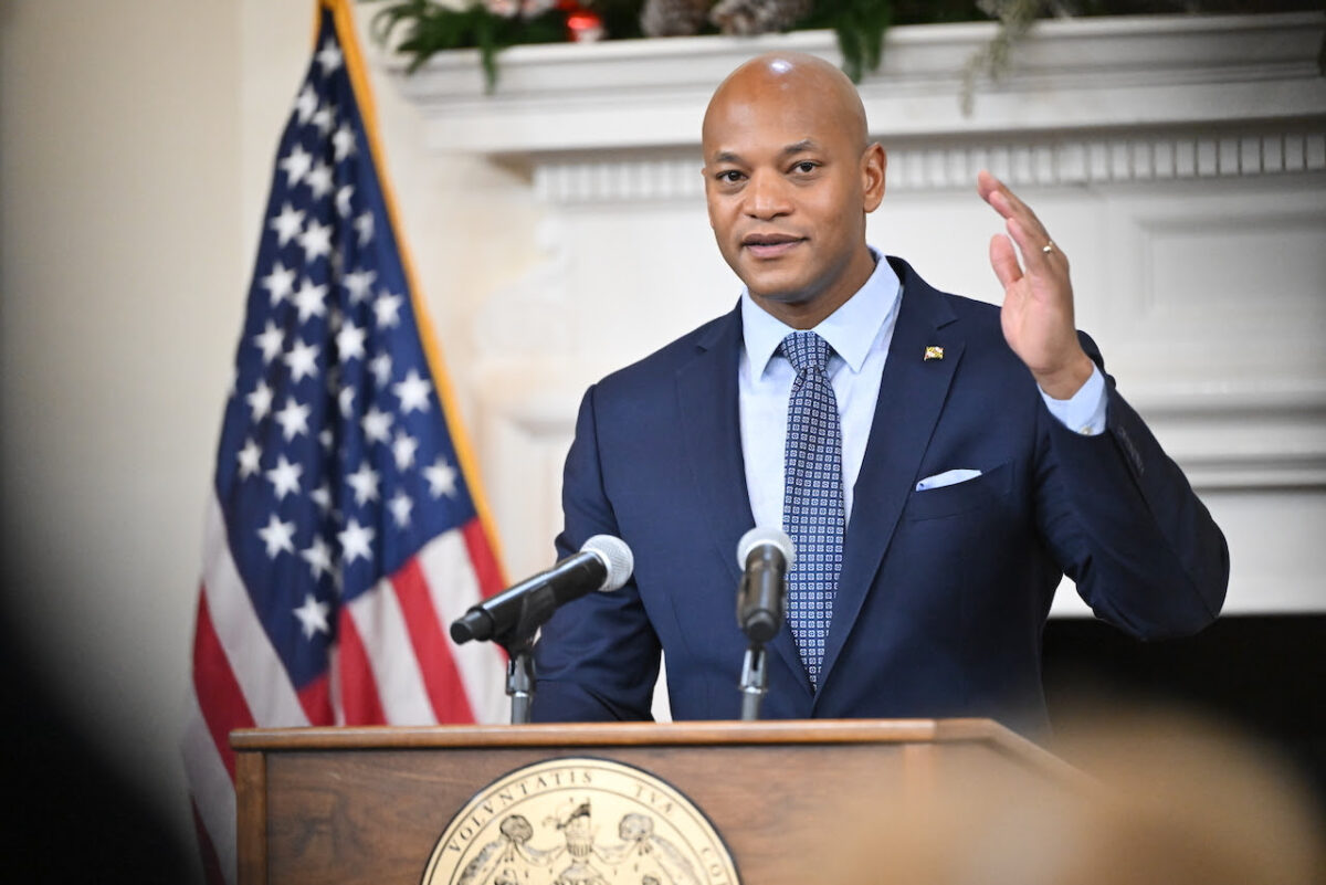 Man in a suit speaking at a podium with microphones beside an American flag.