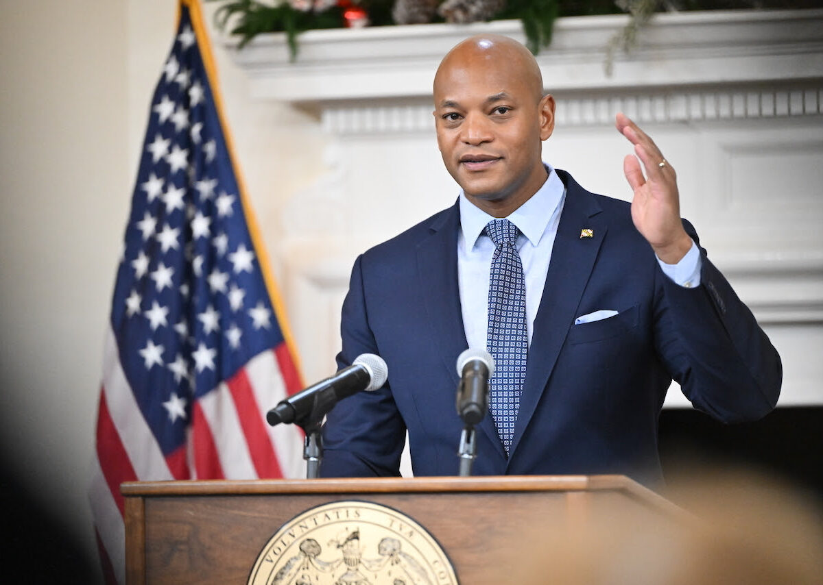 Man in a suit speaking at a podium with microphones beside an American flag.