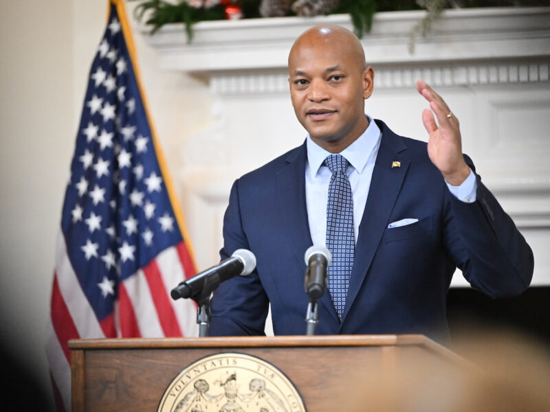 Man in a suit speaking at a podium with microphones beside an American flag.