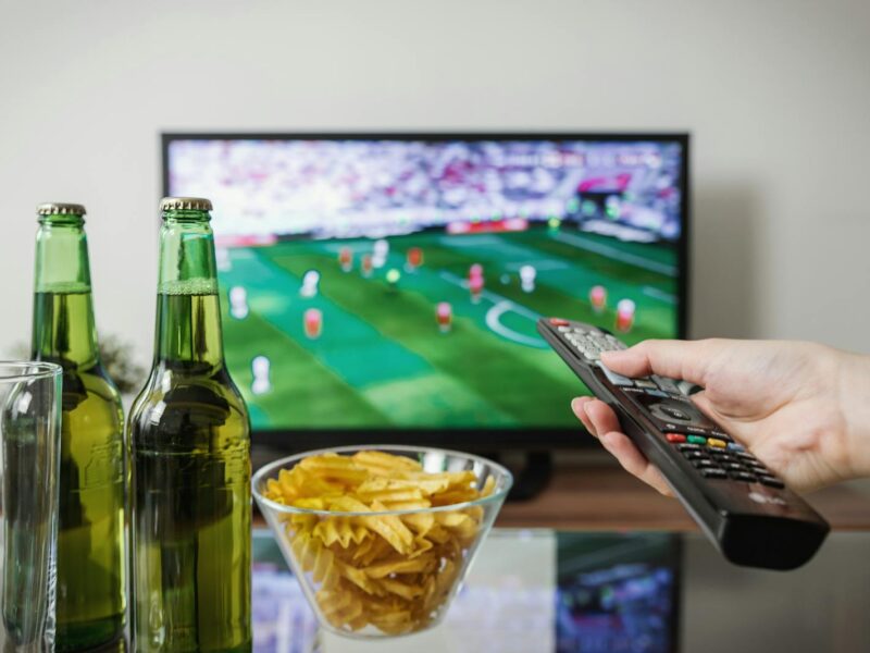 A person holding a remote while watching soccer on TV with beer and snacks on the table.