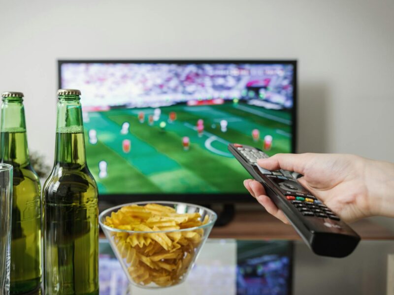 A person holding a remote while watching soccer on TV with beer and snacks on the table.