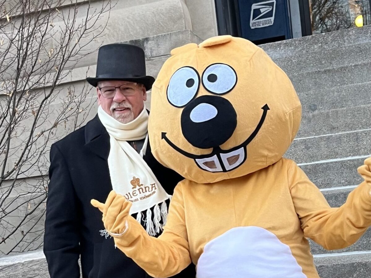 Man in top hat and scarf posing beside a smiling yellow mascot on steps