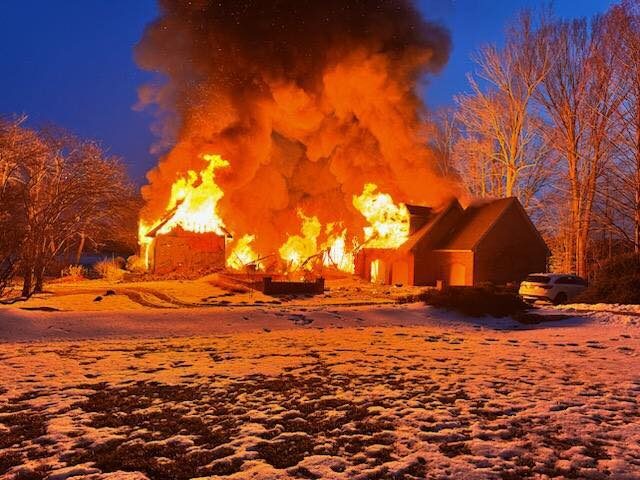 Large house fire engulfing a snowy rural home at dusk with thick black smoke and flames.