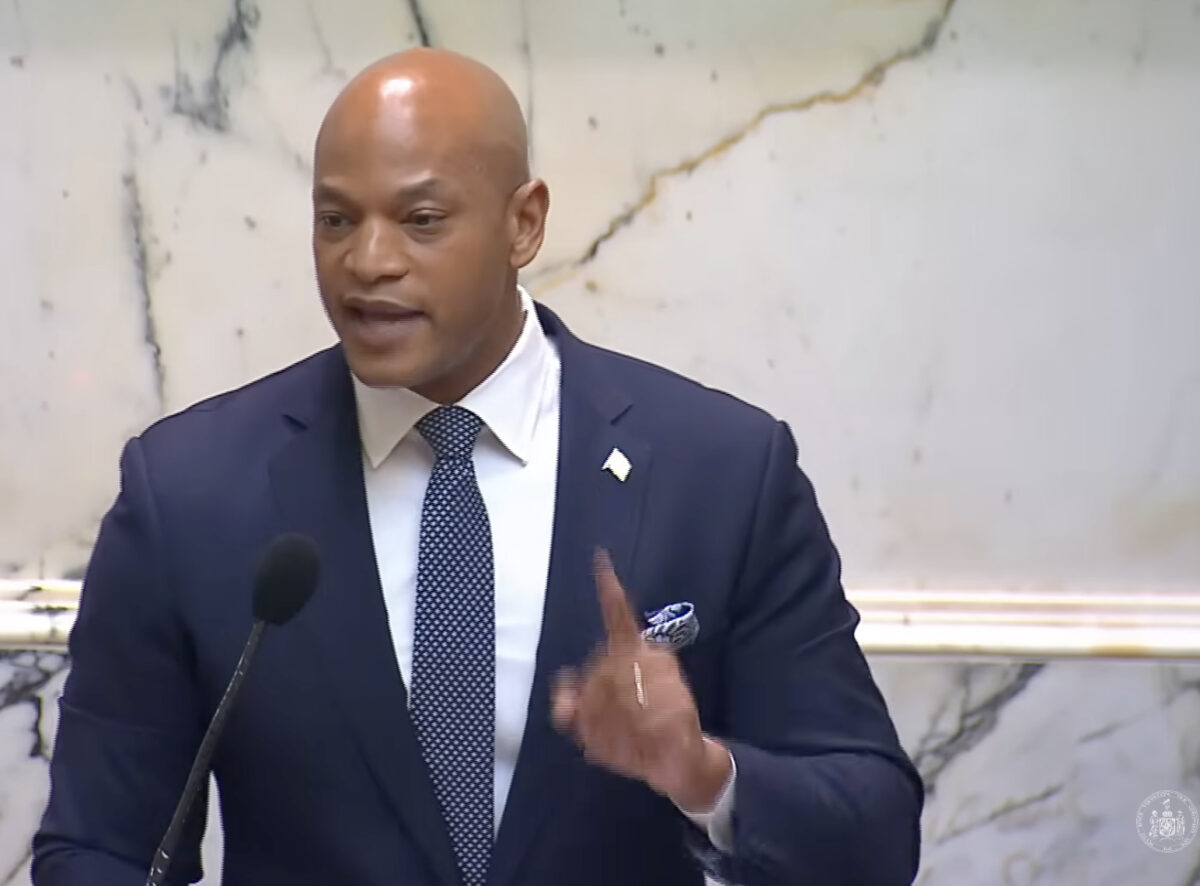 Man in a suit speaking at a podium with a microphone in a marble interior.