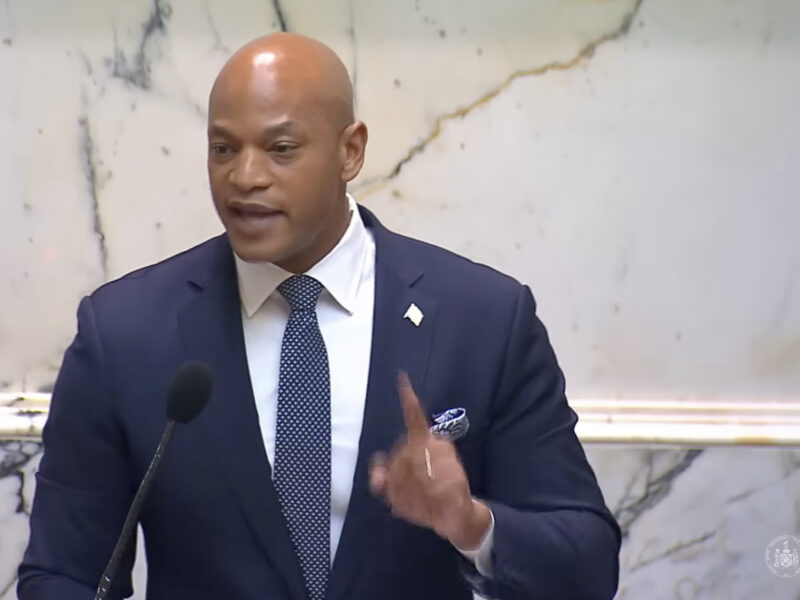 Man in a suit speaking at a podium with a microphone in a marble interior.
