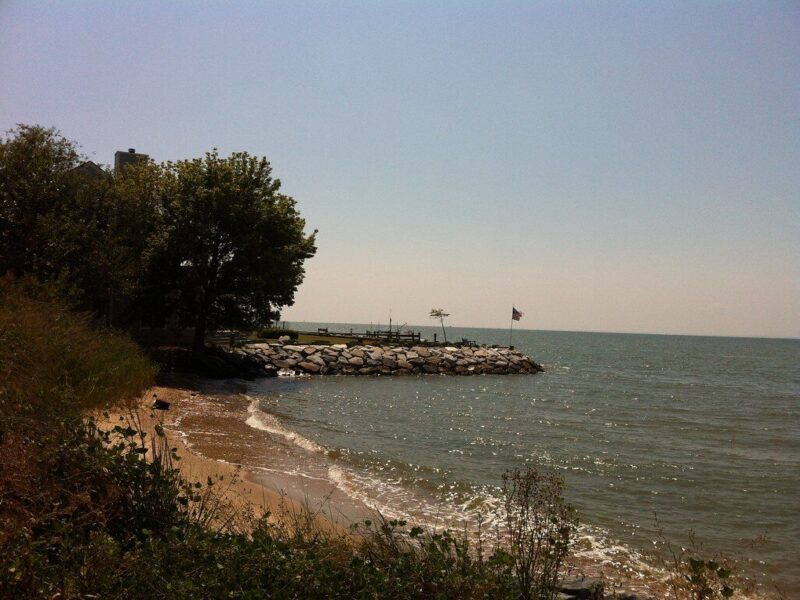 Rocky jetty extends into a calm lake beside a small sandy beach and trees under a clear sky.