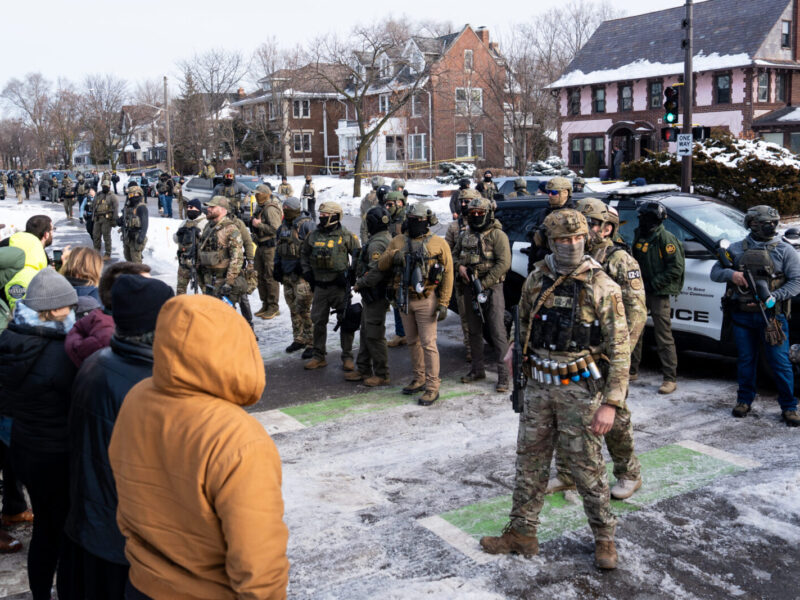 Group of armored police officers stand facing a crowd on a snowy residential street.