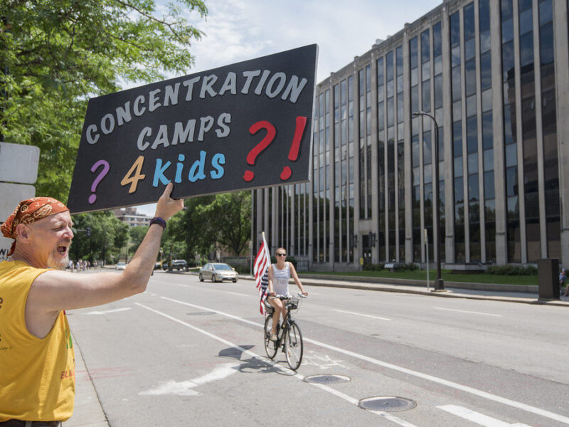 Protester holds a sign reading "Concentration Camps? 4 kids?!" on a city street beside an office building.